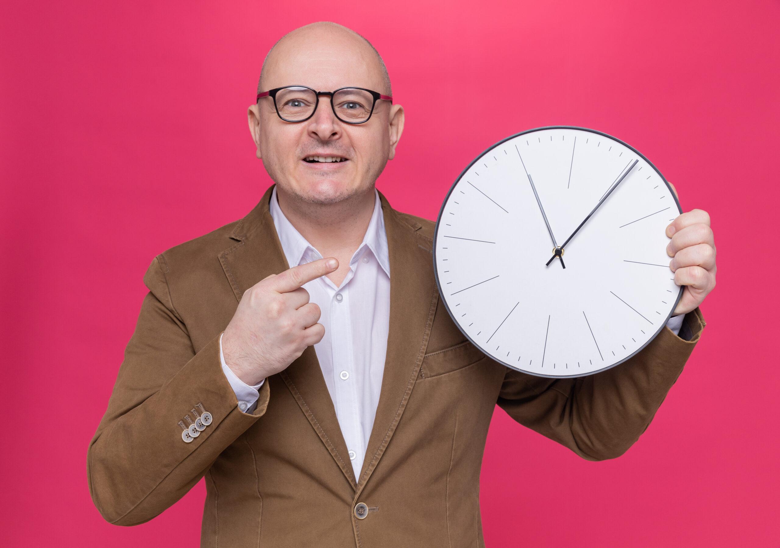 The Aging Clock: How Long You'll Really Live 13 happy middle-aged bald man in suit wearing glasses holding wall clock pointing with index finger at it smiling cheerfully standing over pink background